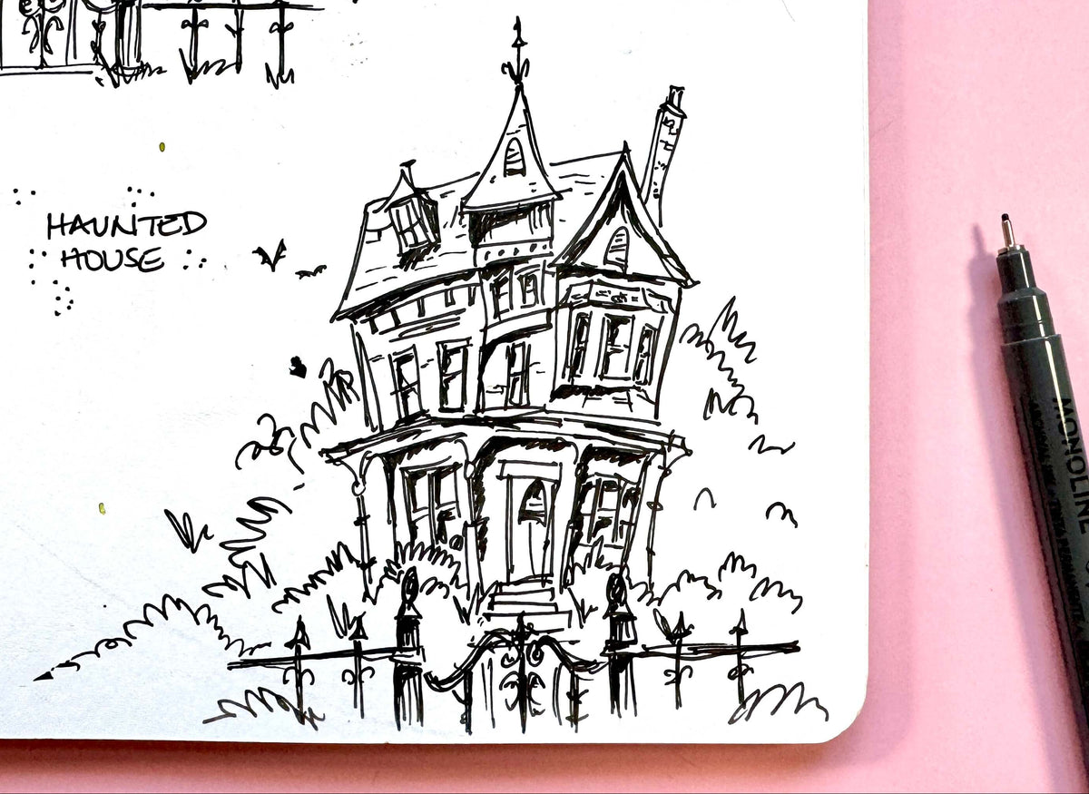 A black ink sketch of a haunted house in a small notebook on a pink background, with a black pen visible on the right side.