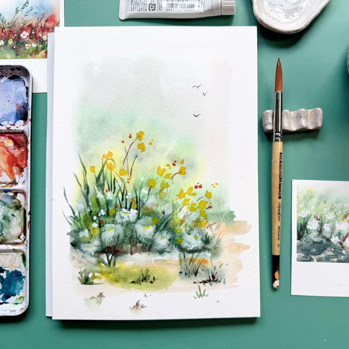 Loose misty watercolor wildflower painting surrounded by a palette, brush, and small studies on a teal table.